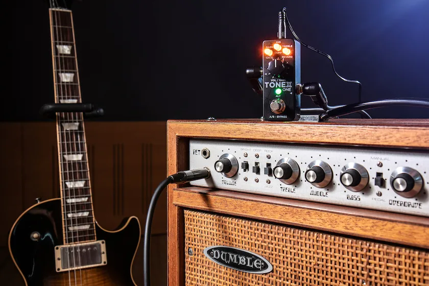 TONEX ONE compact pedal with illuminated display on wooden amp top, black guitar visible left