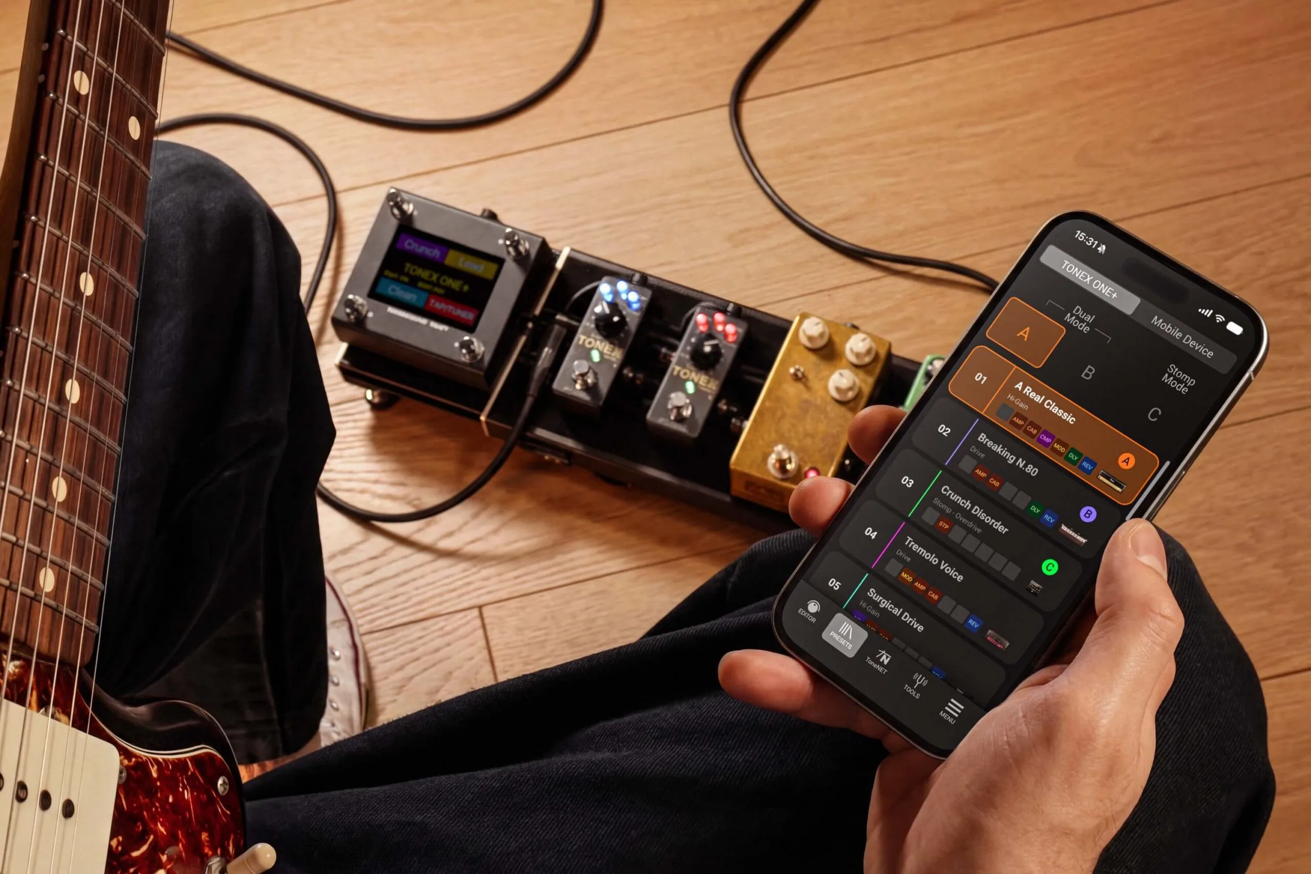 Guitarist holding smartphone displaying TONEX ONE+ app interface, with pedalboard and guitar visible on wooden floor