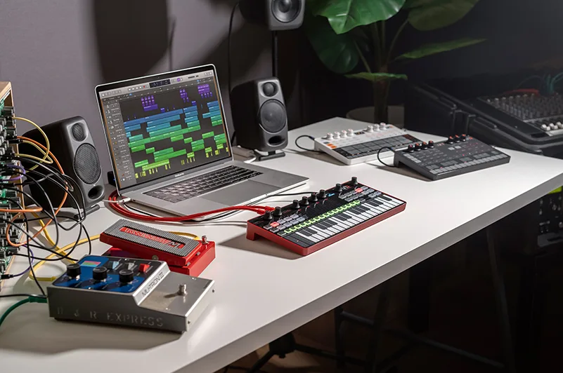Red Uno Synth Pro synthesizer on white desk with laptop displaying DAW, studio monitors, and cables in professional music production setup