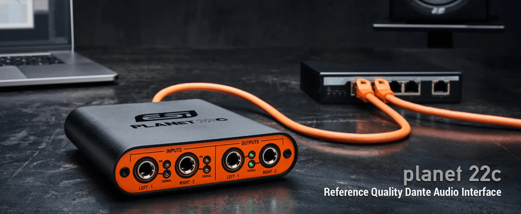ESI Planet 22c Dante audio interface with orange I/O connectors and cable, displayed on dark desk near laptop