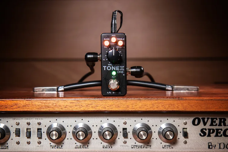 TONEX ONE compact guitar pedal on wooden surface with cables and vintage amplifier control panel below