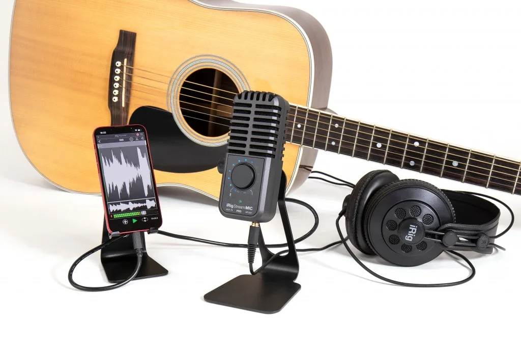 iRig Stream Mic Pro desktop microphone with acoustic guitar, smartphone displaying waveform, and headphones on white background