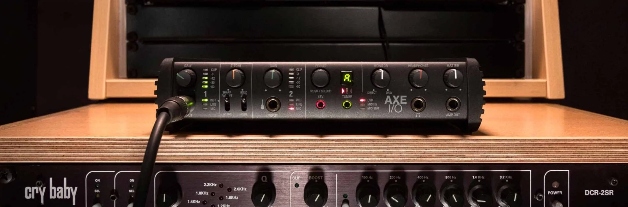 AXE I/O audio interface with black chassis and green LED indicators, positioned on wooden desk above Cry Baby pedal